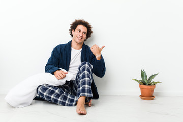 Mature man wearing pajama sitting on house floor points with thumb finger away, laughing and carefree.