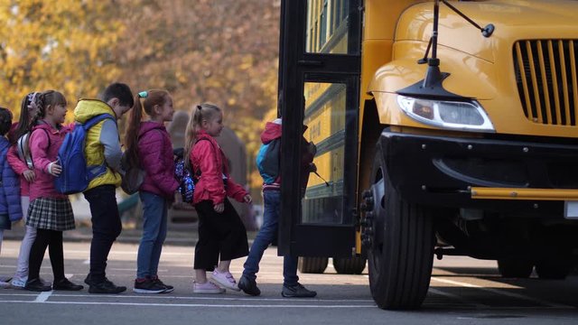 Cute schoolchildren standing in line waving hands while greeting school bus driver and entering transport to go home after studies. Elementary age kids waiting for school bus doors to open outdoors