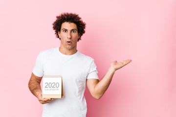 Young caucasian curly man holding a 2020 calendar impressed holding copy space on palm.