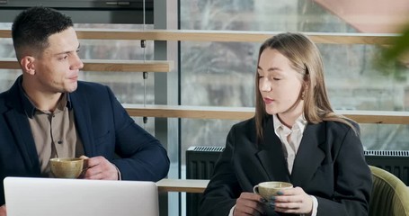 Man and a woman discussing work in the brightly lit modern office. Male and female working with laptop and drink coffee at table. Business people discuss new startup project concept