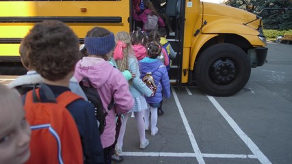 Back view of diverse little students getting on yellow school bus one after another to go home. Preadolescent schoolchildren patiently waiting in line to enter school bus after studies
