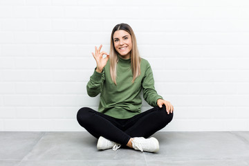 Young caucasian woman sitting on the floor cheerful and confident showing ok gesture.