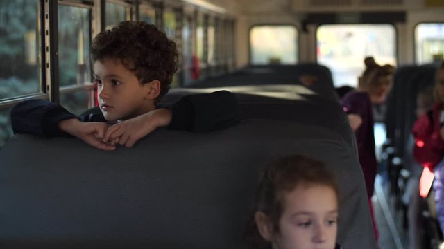 Adorable elementary age boy with curly hair looking out of window while going home on school bus with other students. Diverse preadolescent schoolkids riding school bus after lessons on sunny day