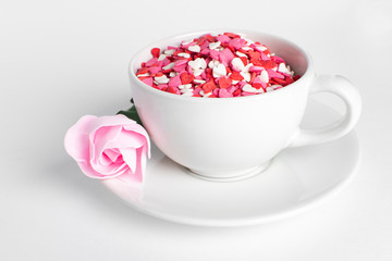 white cup of coffee on a white background filled with heart shaped confetti. pink rose lies on the saucer selective focus. valentine day concept.