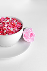 white cup of coffee on a white background filled with heart shaped confetti. pink rose lies on the saucer selective focus. valentine day concept.