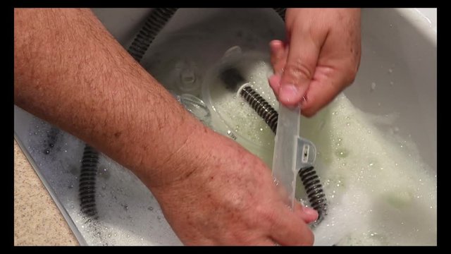 Male Hands Cleaning CPAP Equipment In A Sink Of Soapy Water. Raising The Pieces Out Of The Water To Clean.