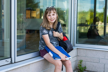 Happy/smiling teen girl/student sitting on a window ledge of her school while holding binders and wearing a backpack.