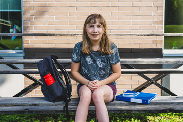 Happy/smiling teen girl sitting on a set of bleachers next to her school with a backpack and binders.