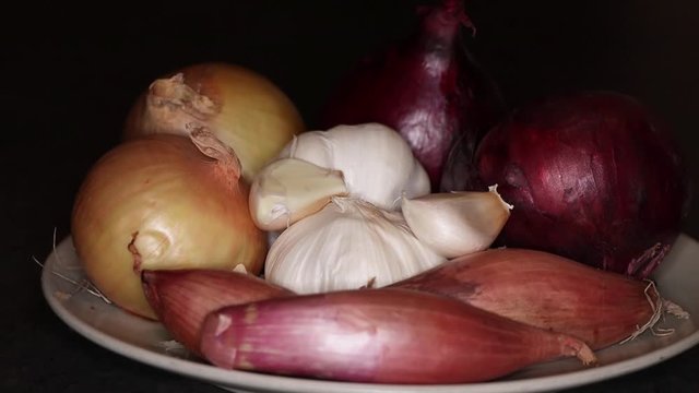 Slow pan of onions, shallots and garlic on a plate with dark background, close-up