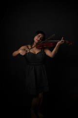 Fashion portrait of an young and attractive Indian Bengali brunette girl with black western dress is playing violin in front of a black studio background. Indian fashion portrait and lifestyle.