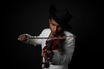 Portrait of young and attractive Indian Bengali brunette man with white western suit and hat is playing violin in front of black studio copy space background. Indian fashion portrait and lifestyle. © abir