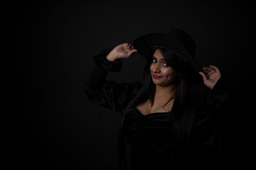 Fashion portrait of an young and attractive Indian Bengali brunette girl with black western dress with a cowboy hat in front of a black studio background. Indian fashion portrait and lifestyle.