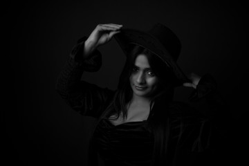 Monochrome portrait of an young and attractive Indian Bengali brunette girl with black western dress with a cowboy hat in front of a black studio background. Indian fashion portrait and lifestyle.