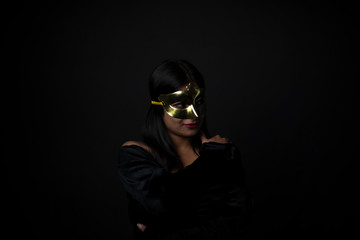 Fashion portrait of an young and attractive Indian Bengali brunette girl with black western dress with golden mask in front of a black studio background. Indian fashion portrait and lifestyle.