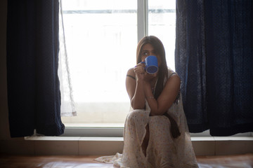 An young and attractive Indian  brunette woman in white sleeping wear drinking coffee near the window in white background. Indian lifestyle.