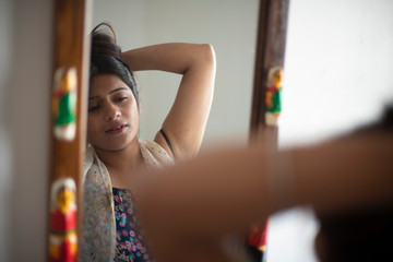 Reflection of an young and attractive Indian Bengali brunette woman is looking her face in a mirror while tying her hair in the morning in her room. Indian lifestyle.