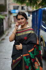 Fototapeta premium Portrait of a beautiful brunette Indian bengali thoughtful woman in traditional wear sari standing in a lane on a drenched morning of Durga Puja festival in urban background. Indian lifestyle.