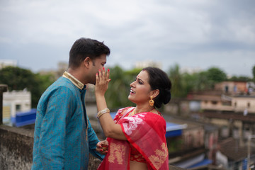 A brunette Indian Bengali romantic couple in traditional wear interacting between themselves on a roof top in romantic mood in the morning of Durga Puja festival in urban background. Indian lifestyle.