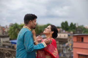 A brunette Indian Bengali romantic couple in traditional wear interacting between themselves on a roof top in romantic mood in the morning of Durga Puja festival in urban background. Indian lifestyle.