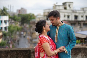 A brunette Indian Bengali romantic couple in traditional wear interacting between themselves on a roof top in romantic mood in the morning of Durga Puja festival in urban background. Indian lifestyle.