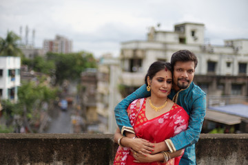 A brunette Indian Bengali romantic couple in traditional wear interacting between themselves on a roof top in romantic mood in the morning of Durga Puja festival in urban background. Indian lifestyle.