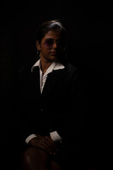 An young and attractive dark skinned Indian Bengali brunette woman in western jacket, skirt and goggles sitting under spotlight in a black copy space background. Indian fashion photography.