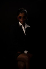 An young and attractive dark skinned Indian Bengali brunette woman in western jacket, skirt and goggles sitting under spotlight in a black copy space background. Indian fashion photography.