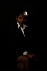 Fototapeta na wymiar An young and attractive dark skinned Indian Bengali brunette woman in western jacket and skirt sitting under spotlight in a black copy space background. Indian fashion photography.