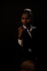 Fototapeta premium An young and attractive dark skinned Indian Bengali brunette woman in western jacket, skirt and goggles sitting under spotlight in a black copy space background. Indian fashion photography.