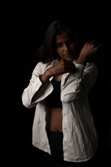 Fashion portrait of young and attractive dark skinned Indian Bengali brunette woman in western lingerie and glasses removing white shirt  in black copy space background. Indian fashion photography.