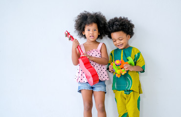 Two young African brother and sister  stand in front of white wall and girl holds small guitar and boy holds some toys in hand with day light.