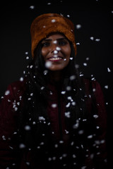 Portrait of an young and attractive dark skinned Indian woman wearing woolen cap and jacket playing with snowflakes in a snowy evening in dark background. Winter and Christmas