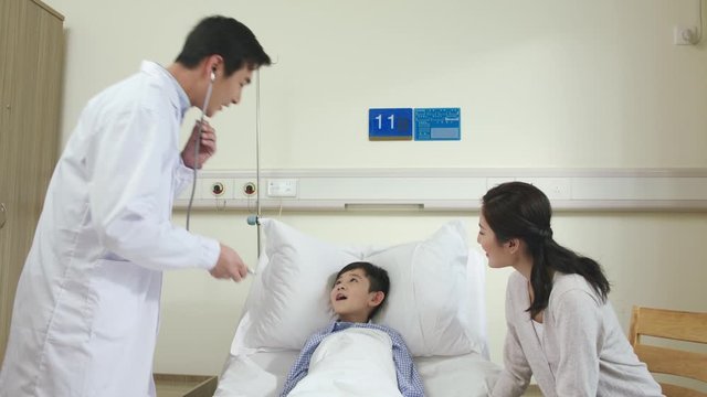 young asian male doctor making rounds examining and talking to child patient and mother in hospital ward