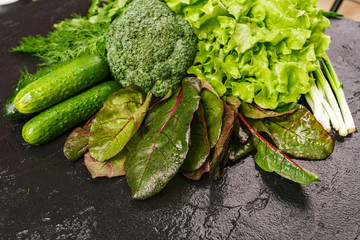 A mixture of fresh produce on a dark table. Dill, green pepper, parsley, cucumbers, broccoli, onions, borax leaves.