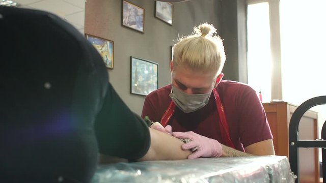 Master doing tattooing in tattoo studio. Professional tattooist works in studio. A man in pink gloves makes a tattoo on the leg of a young girl. Slow motion.