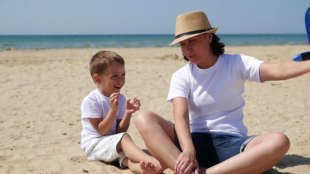 Happy family: a mother and her son are sitting on a sandy beach on a sunny summer day and playing with a toy airplane