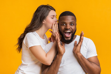 Young woman sharing secret with her shocked black boyfriend