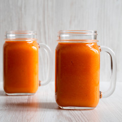 Homemade Mango Carrot Smoothie in glass jar mugs over white wooden background, side view.