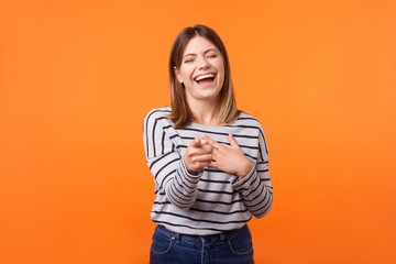 Fototapeta premium Portrait of amusing joyous young woman with brown hair in long sleeve striped shirt closing eyes, pointing finger and laughing out loud at funny joke. indoor studio shot isolated on orange background