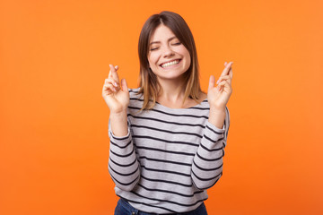 Portrait of hopeful joyous young woman with brown hair in long sleeve shirt standing, raising finger crossed while making wish, confident to win. indoor studio shot isolated on orange background
