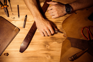 Working process of the leather belt in the leather workshop. Man holding hands on wooden table. Crafting tools on background. Tanner in old tannery. Close up men arm.
