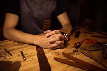 Working process of the leather belt in the leather workshop. Man holding hands on wooden table. Crafting tools on background. Tanner in old tannery. Close up men arm. Interlock fingers.