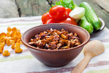 Fried chanterelles with onion in rustic bowl and plate with fresh vegetables for salad on background