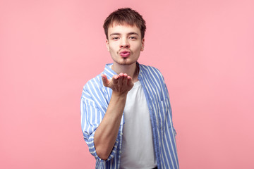 Portrait of handsome amorous brown-haired man with small beard and mustache in casual striped shirt holding palm up and sending air kiss, flirting. indoor studio shot isolated on pink background