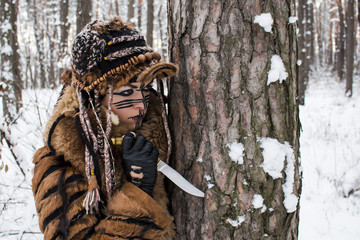 A girl hunter in fur clothes holds a knife stuck in the bark of a tree, because of which she tracks...
