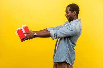 Take this present! Side view of generous man in denim casual shirt holding out red gift box and smiling, congratulating on holidays, giving surprise. indoor studio shot isolated on yellow background
