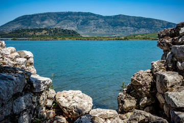 Fototapeta premium Sunny spring day view of salt lagoon lake, valley and mountain in the national park of Butrint in Albania, Europe. Fortress stone ruined wall in the foreground.