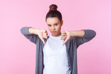 Dislike! Portrait of dissatisfied brunette teenage girl with bun hairstyle in casual clothes making thumbs down gesture, expressing discontent disapproval, critique. studio shot, pink background