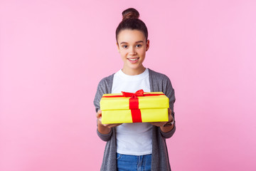 Holiday present. Portrait of positive teenage brunette girl with bun hairstyle in casual clothes holding gift box and smiling at camera, enjoying surprise. studio shot isolated on pink background