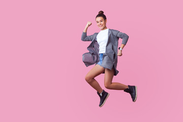 Ecstatic brunette teenage girl with bun hairstyle in casual clothes jumping in the air with...